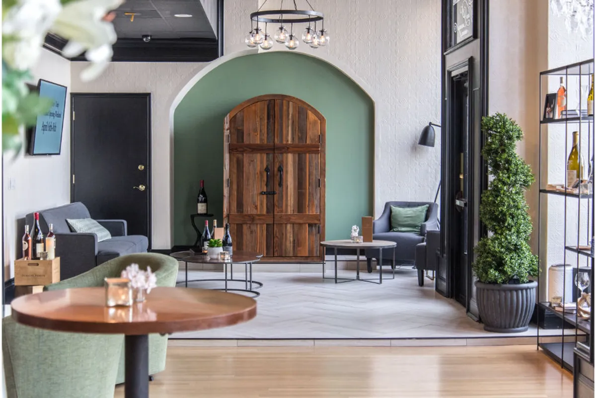 A warm, inviting guest room with a green accent wall, comfortable seating arrangements, a wooden door, and a display shelf stocked with bottles, suggesting the presence of an in-room honor bar. The setting feels both cozy and functional, ideal for relaxation or casual work.