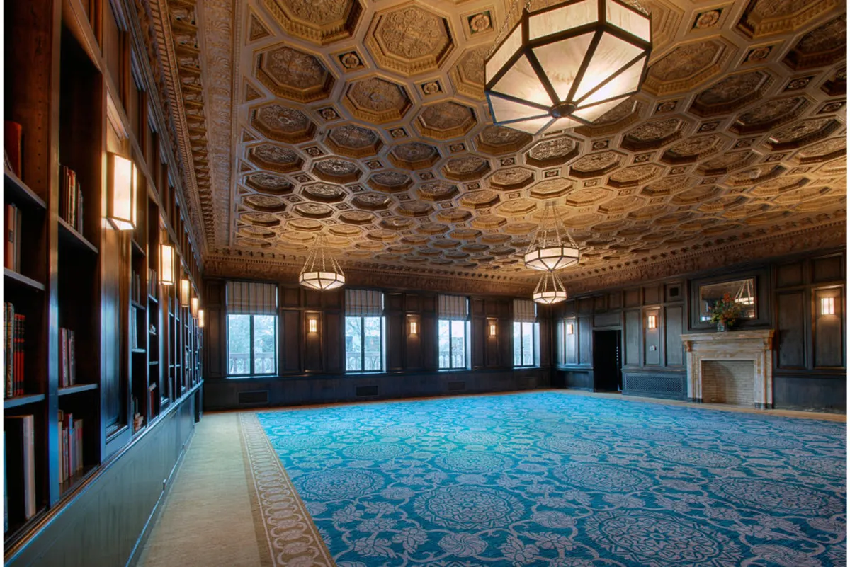 A refined and inviting interior setting with an ornate ceiling, large windows letting in natural light, bookshelves, a fireplace, and an intricate carpet. The space is suited for business meetings, networking events, or informal gatherings, embodying sophistication and productivity.