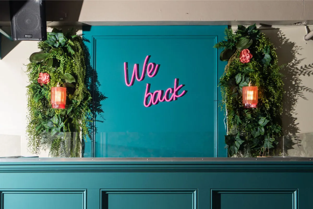 A playful interior scene with a neon sign reading 'We back', vibrant floral arrangements, lush greenery, and a cheerful ambiance. Designed for socializing and relaxation, it serves as a unique photo backdrop and gathering spot.