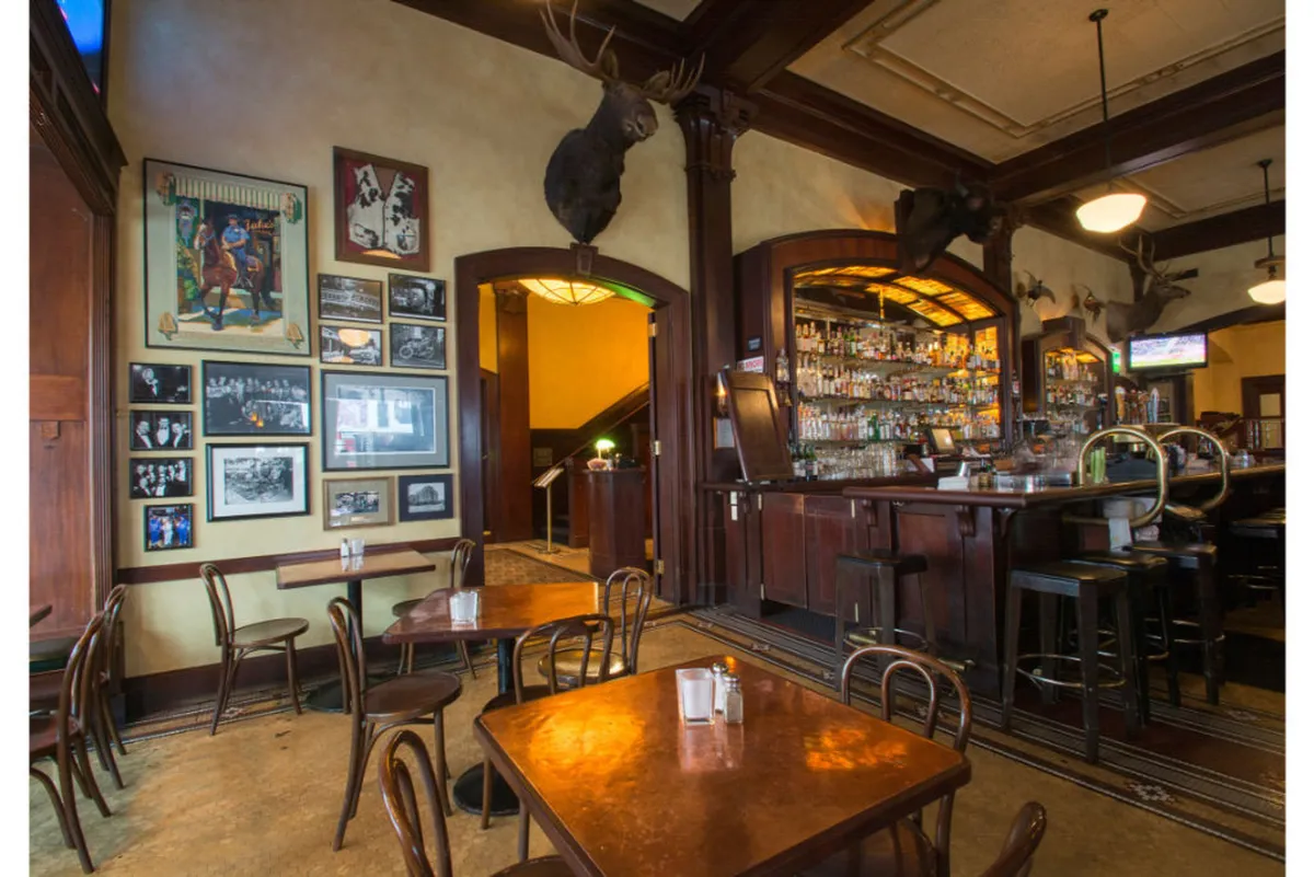 A casual and relaxed bar area featuring wooden tables and chairs, shelves stocked with a variety of liquors, framed photographs on the wall, and quirky moose heads mounted above. The environment suggests sociability and a welcoming public space where guests can unwind.