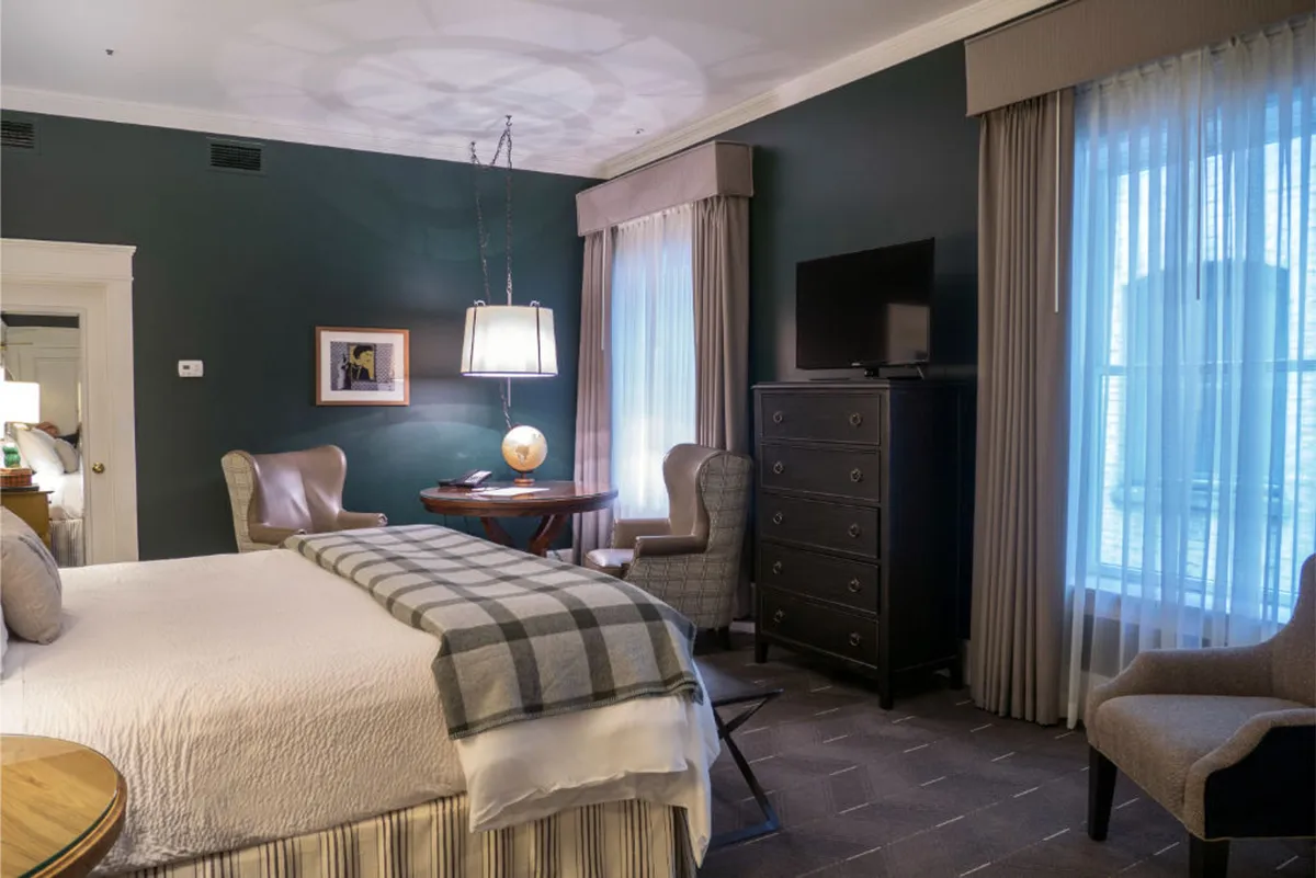 This inviting hotel room features a plush, neatly dressed bed as the central focus, flanked by elegant armchairs and a cozy reading nook by the window. A stylish dresser and a table topped with a classic globe add sophistication, while soft lighting creates a serene, relaxing ambiance ideal for unwinding after exploring Portland.