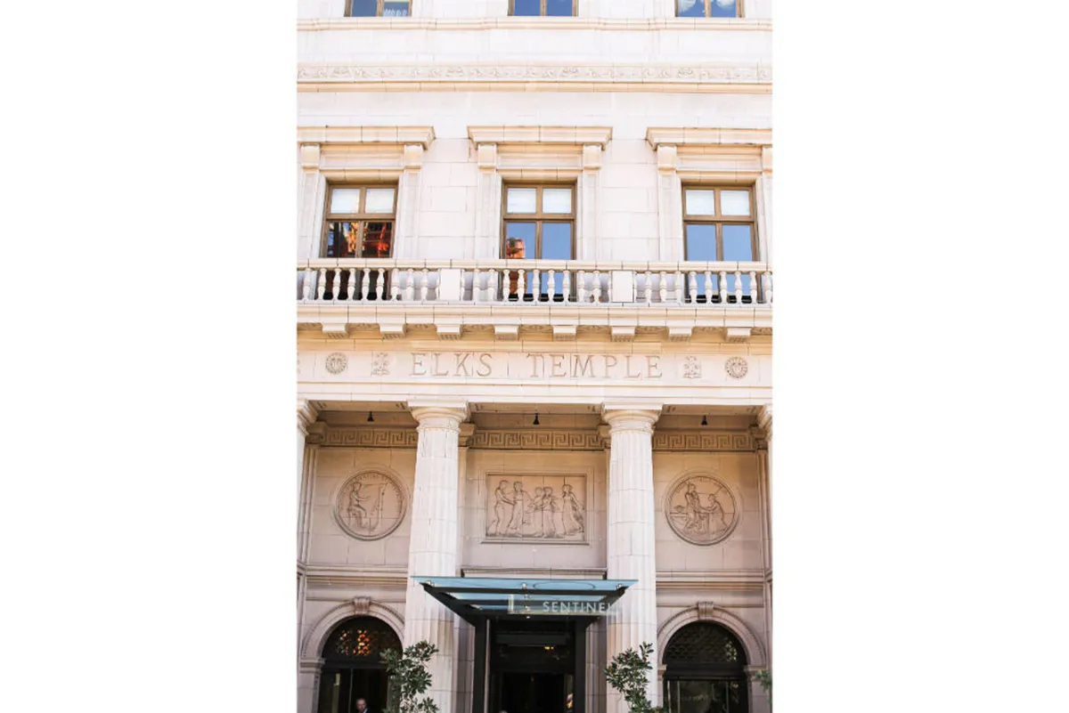 The exterior of the Elks Temple, featuring its distinctive sign, ornate columns, decorative medallions, and expansive windows. The entrance welcomes guests, and the building's historic architecture stands out against its urban surroundings.