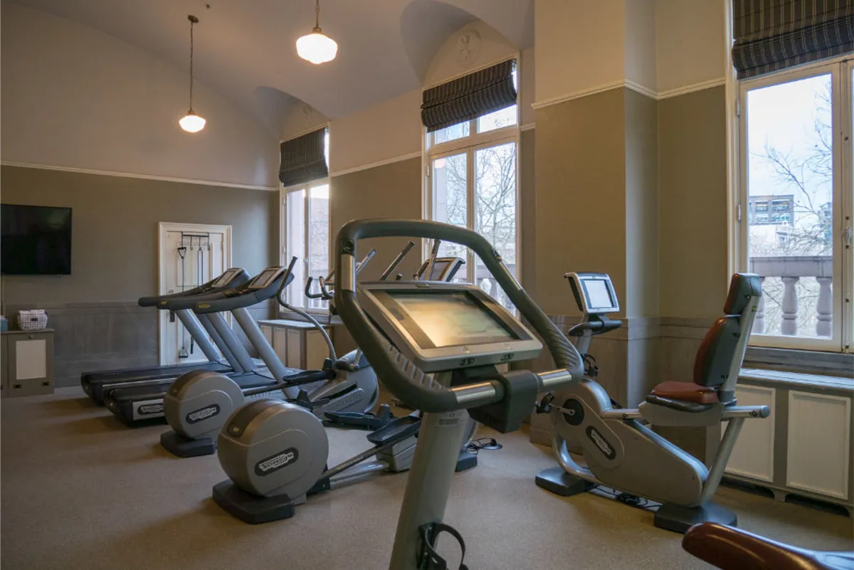 A contemporary fitness center with treadmills, elliptical machines, large windows for natural light, and a TV/monitor. The space is open, bright, and designed for both individual workouts and group classes.