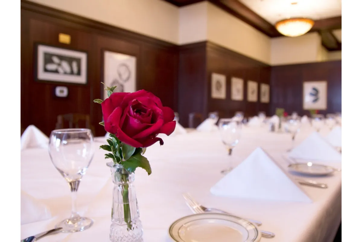 An elegant table setting featuring a single red rose, stylish tableware, and framed artwork in the background. The atmosphere is warm and sophisticated, perfect for fine dining or special celebrations.