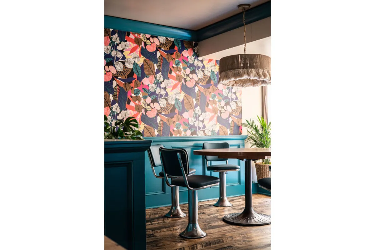 A cheerful space featuring colorful floral wallpaper, a sturdy wooden table, stylish chairs, a hanging light fixture, and lush potted plants. This bright and modern ambiance is perfect for enjoying a cup of Portland Roasters coffee or Smith tea.